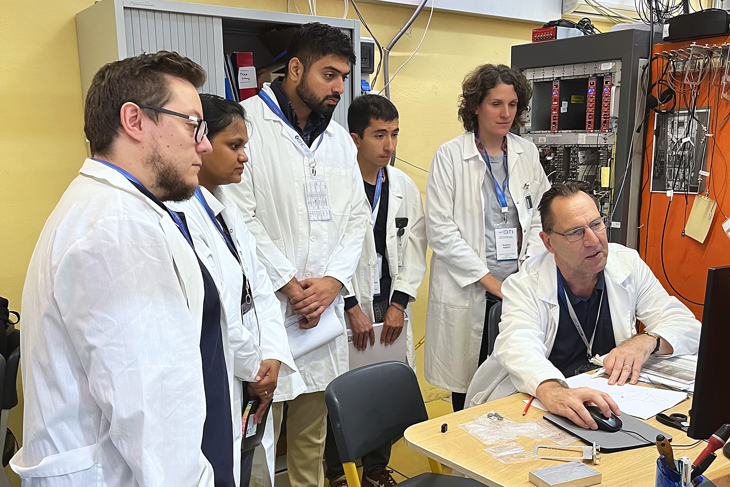 CETS 2026 participants in laboratory coats watching an instructor demonstrate measurement software during a practical session at the Budapest Neutron Centre.