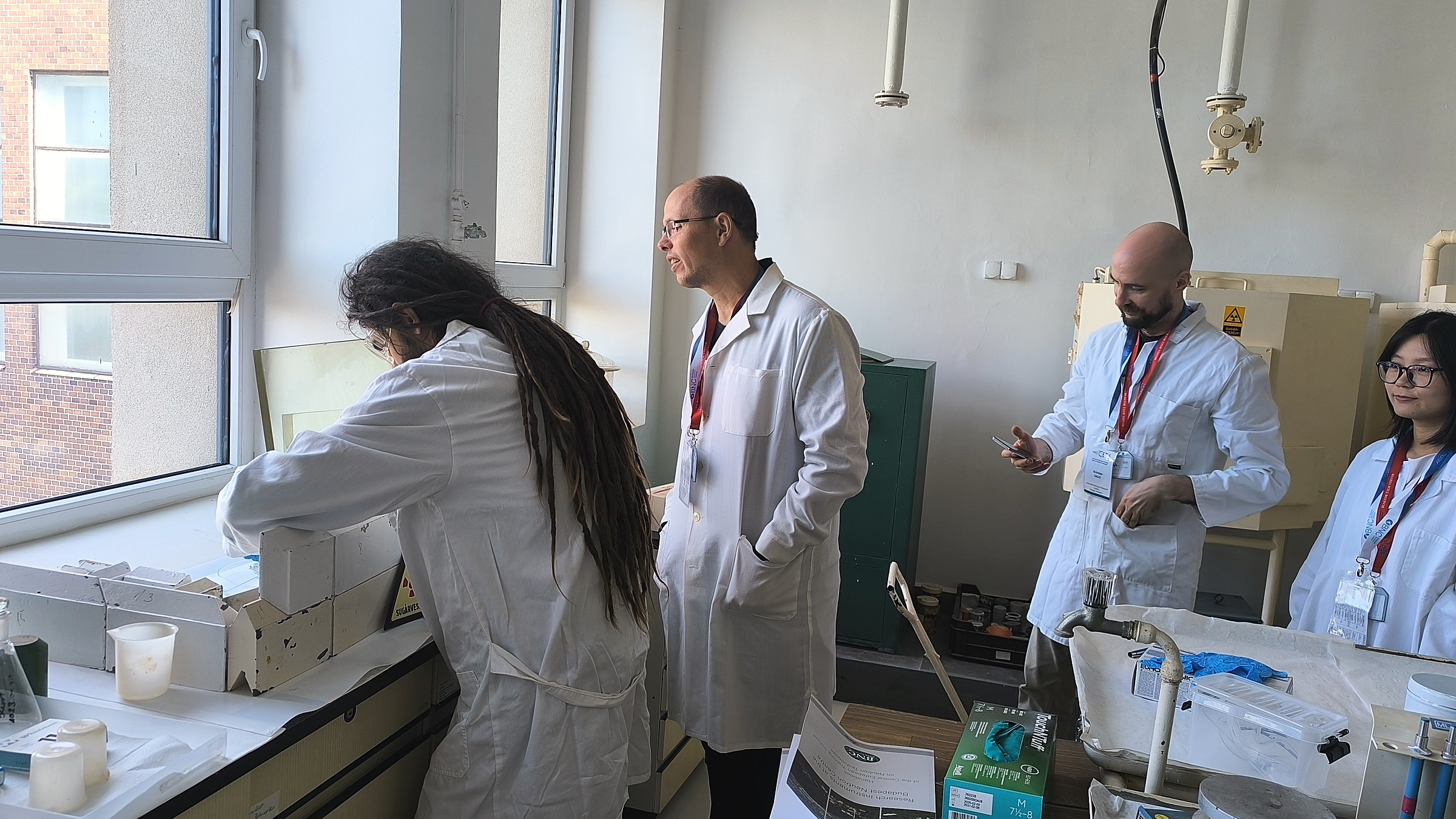 CETS 2026 participants observing the unpacking of a radioactive sample behind protective shielding at the Budapest Neutron Centre.